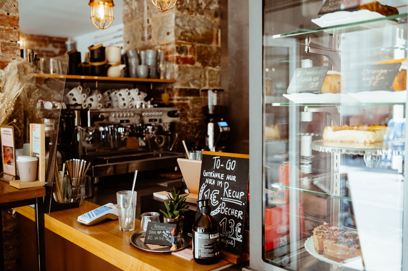 Bild des Verkaufstresens im Café Gustav mit einer Kaffeemaschine und Tassen im Hintergrund, Kuchen in einer Vitrine und einem EC-Gerät zum Bezahlen 