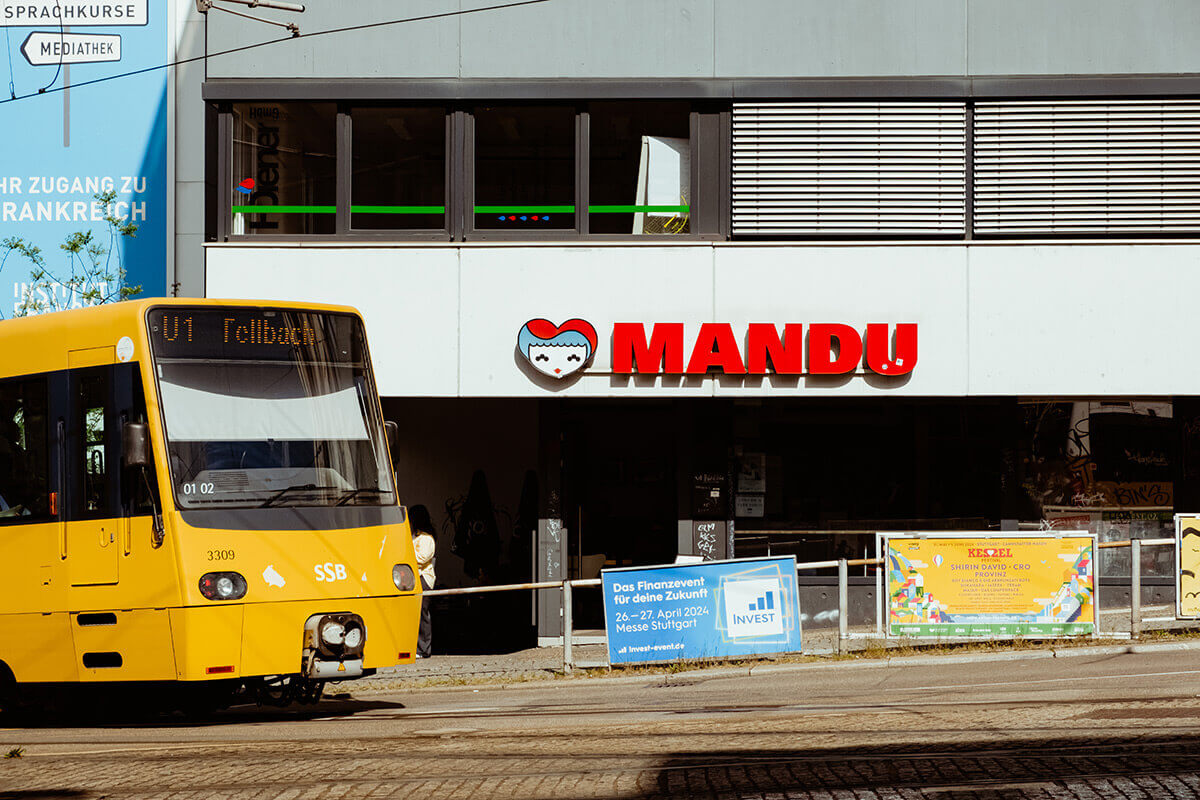Außenansicht des koreanischen Restaurants Mandu, wo gerade die gelbe Stadtbahn der Linie U1 Fellbach der SSB vorbeifährt
